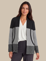 A woman modeling a color-block knit cardigan with black, gray, and white textured panels, worn over a white blouse and dark gray trousers against a solid beige background.