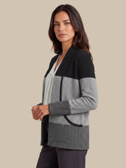 A woman modeling a color-block knit cardigan with black, light gray, and dark gray textured panels, paired with a white top and dark pants, set against a solid beige background.
