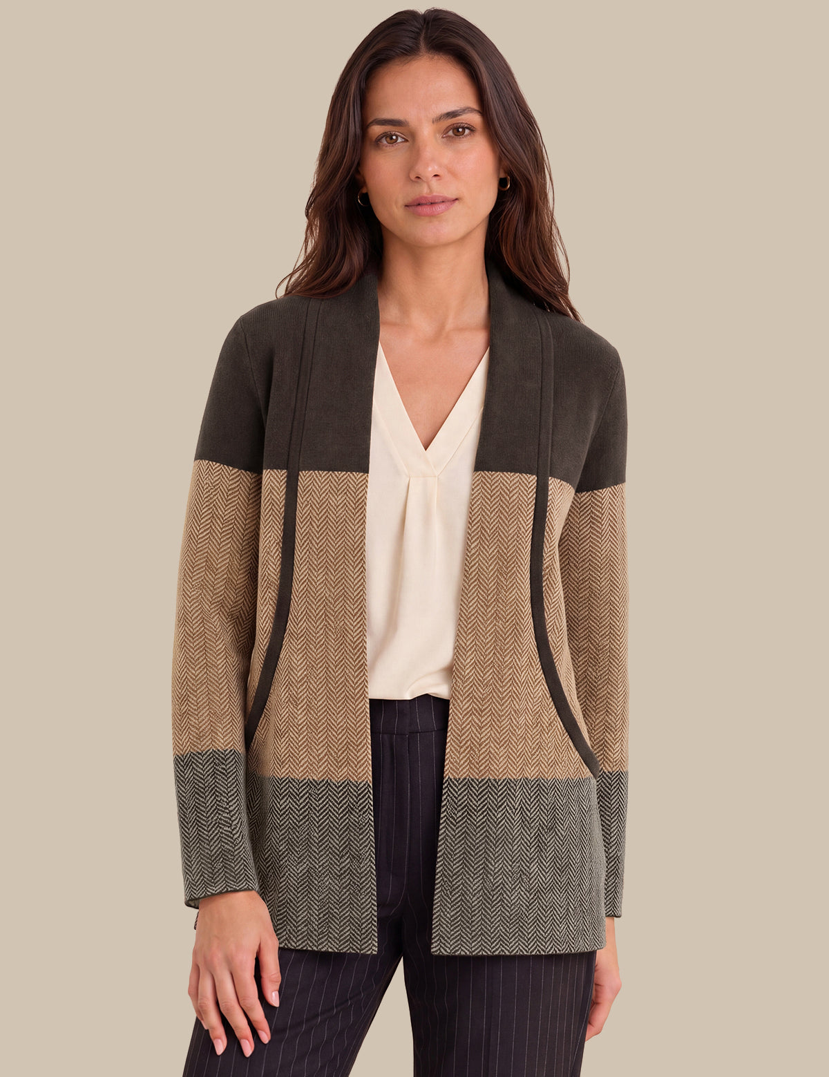 A woman modeling a color-block cardigan with a dark olive green top section, a tan and beige herringbone middle section, and a gray herringbone bottom section, worn over a cream blouse and dark pinstripe trousers against a solid beige background.