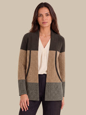 A woman modeling a color-block cardigan with a dark olive green top section, a tan and beige herringbone middle section, and a gray herringbone bottom section, worn over a cream blouse and dark pinstripe trousers against a solid beige background.