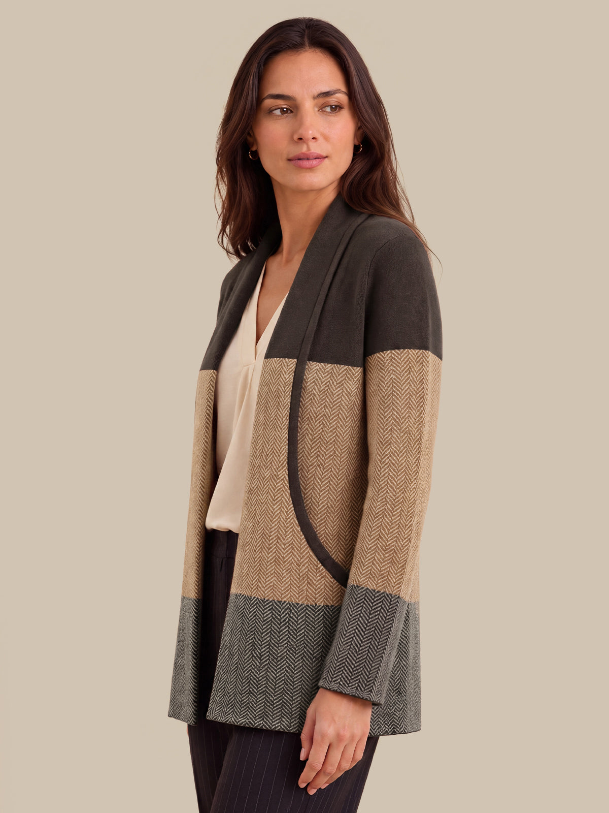 A woman modeling a color-block shawl-collar cardigan with a herringbone pattern. The cardigan features blocks of dark brown, beige, and gray, styled over a cream top against a solid beige background.