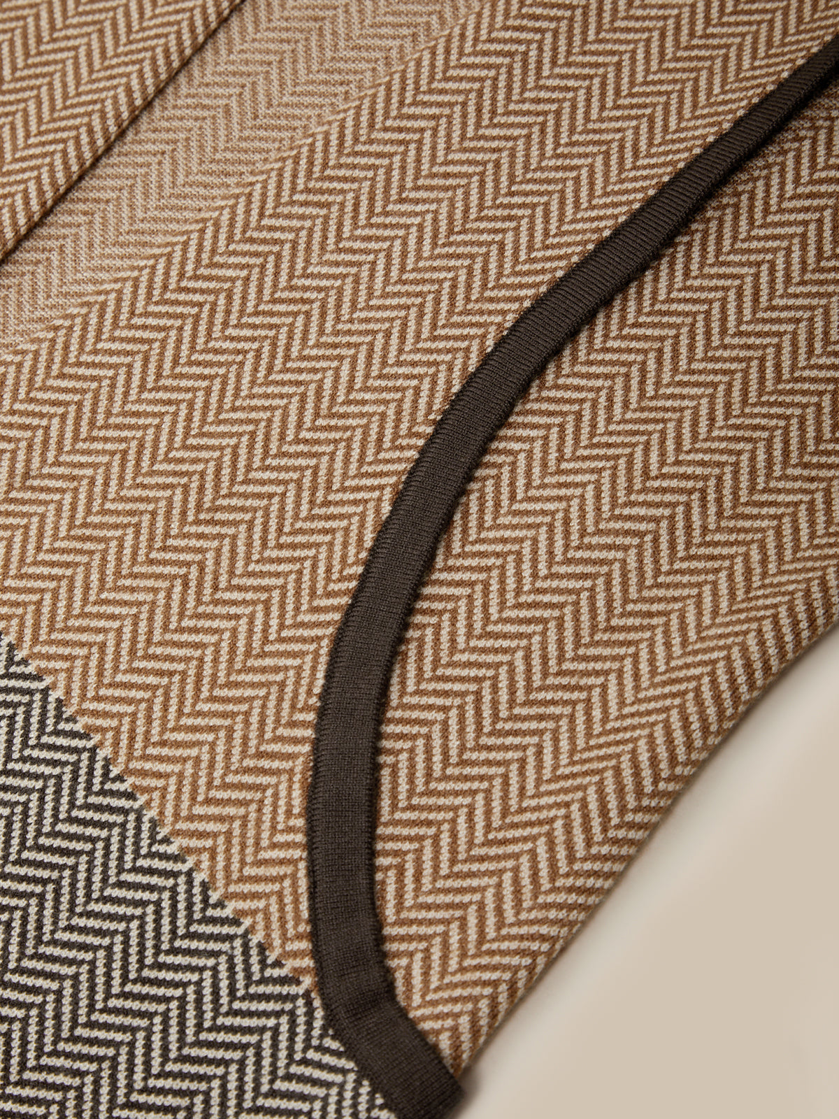Close-up of a woven textile with a tan and brown herringbone pattern, a dark brown curved trim, and a small section of a gray-and-white herringbone pattern at the corner.