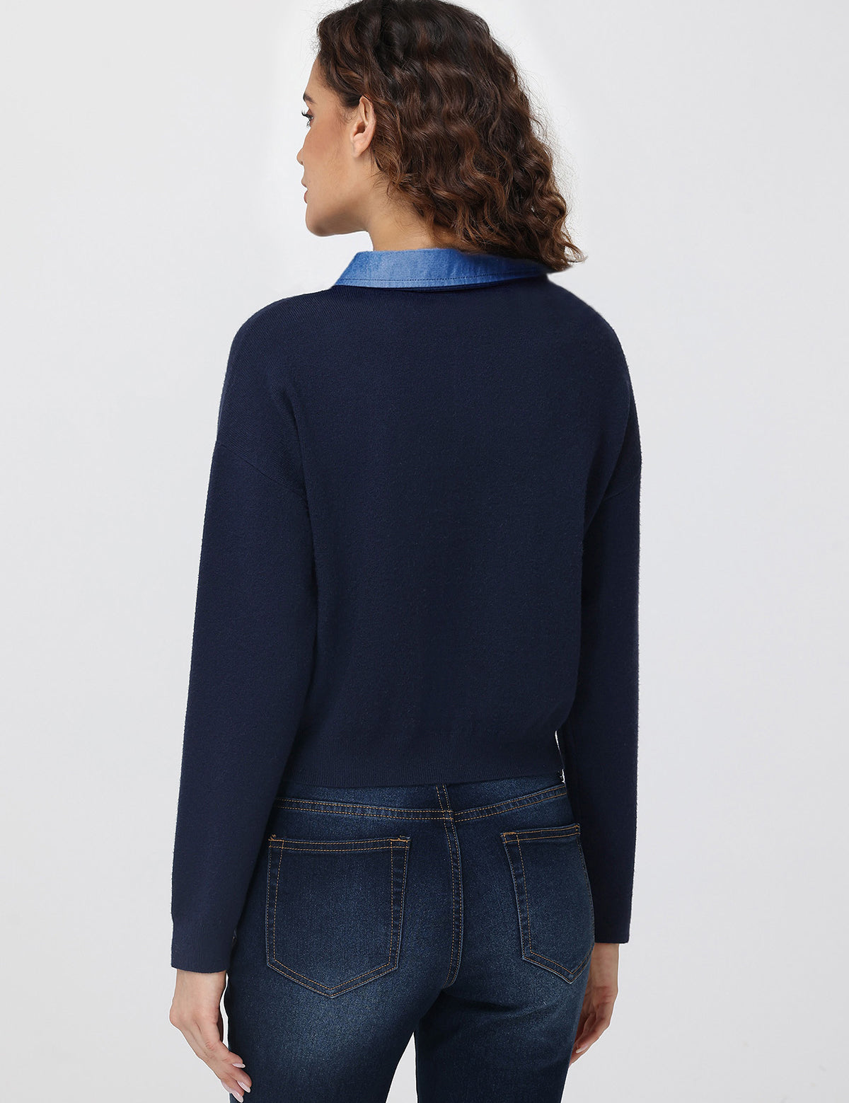 Person wearing a navy sweater with a blue collar and dark jeans on a light gray background