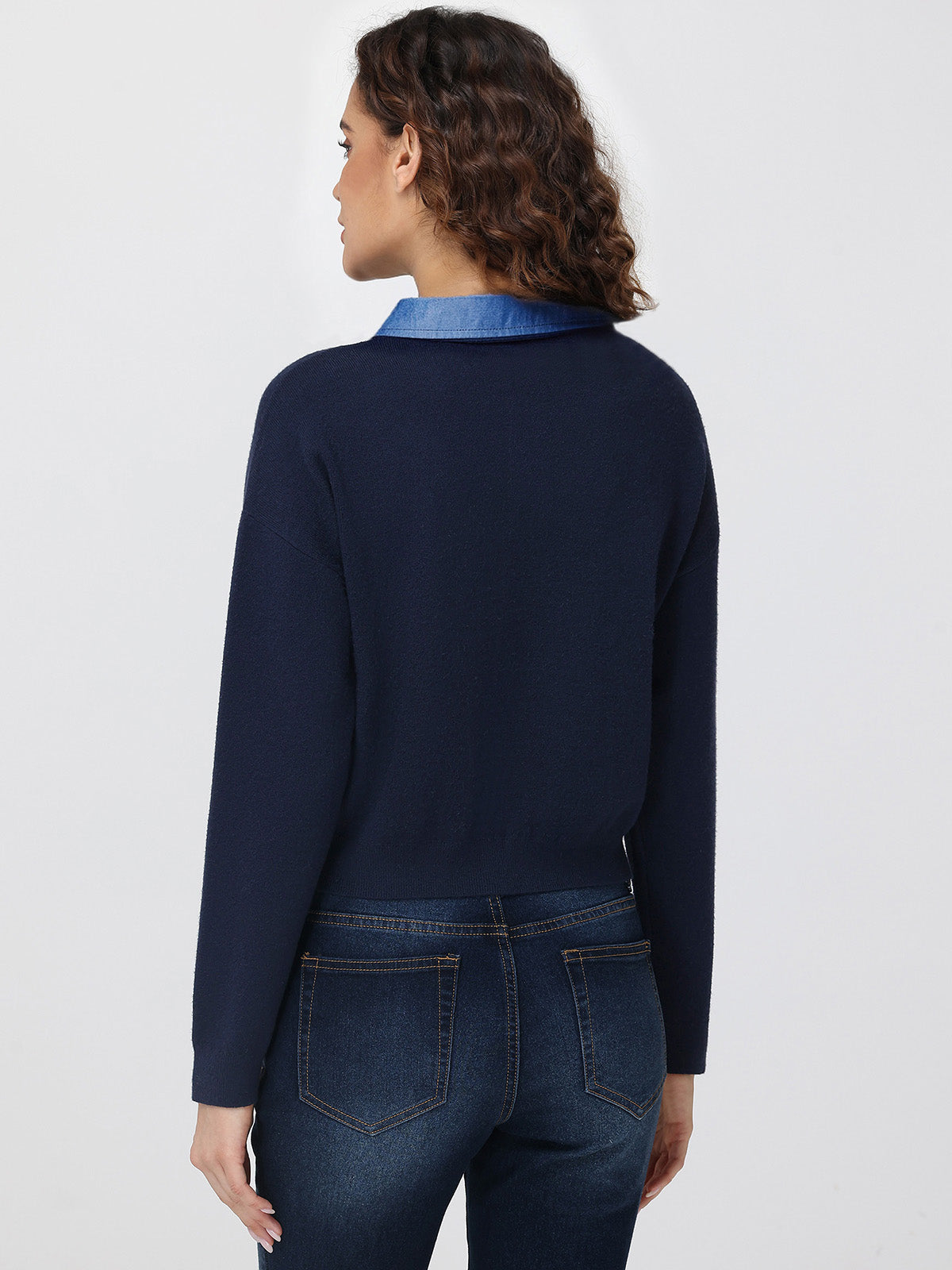 Person wearing a navy sweater with a blue collar and dark jeans on a light gray background