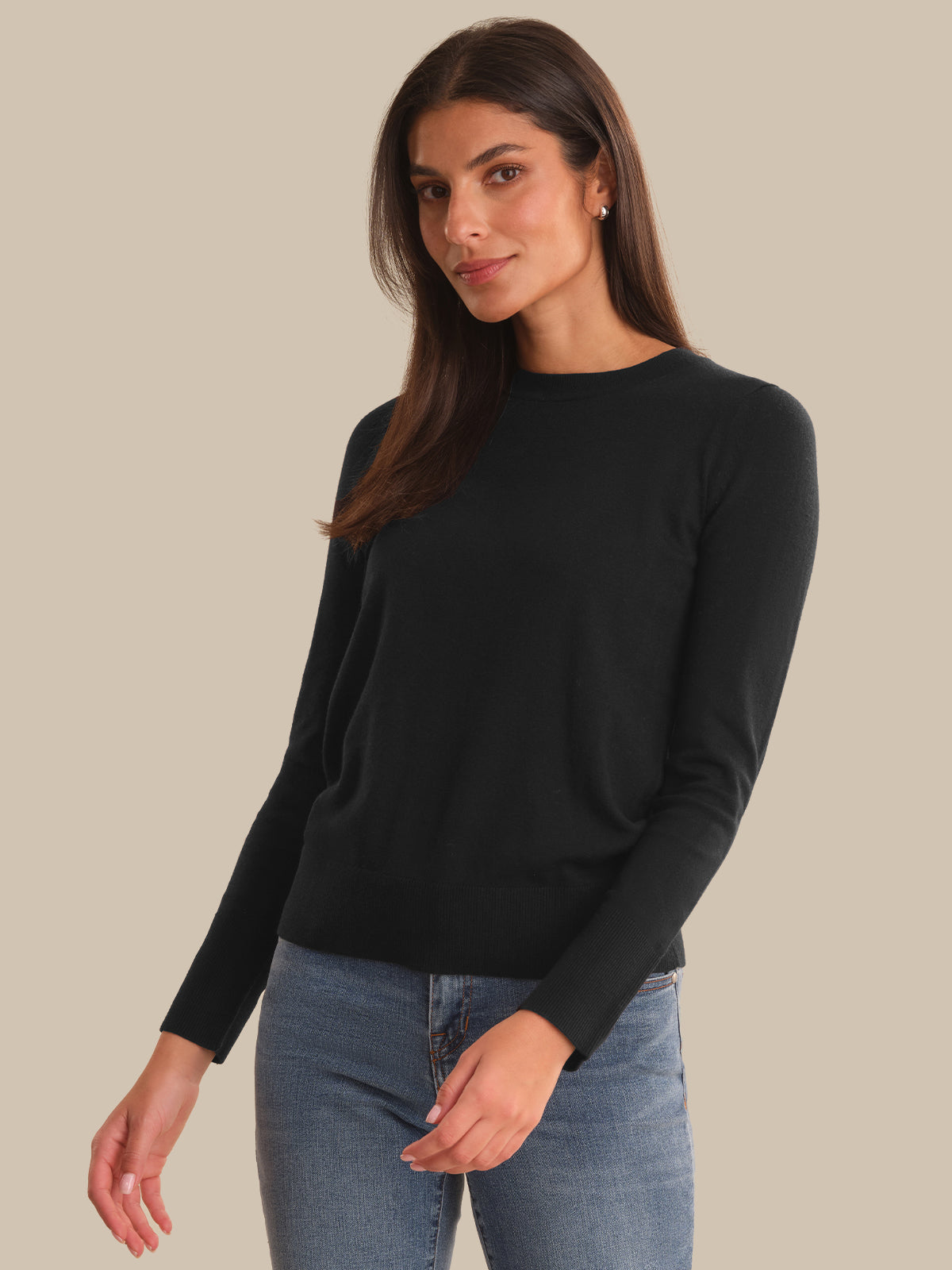 A woman modeling a black long-sleeve crewneck sweater paired with blue jeans, set against a solid beige studio background.