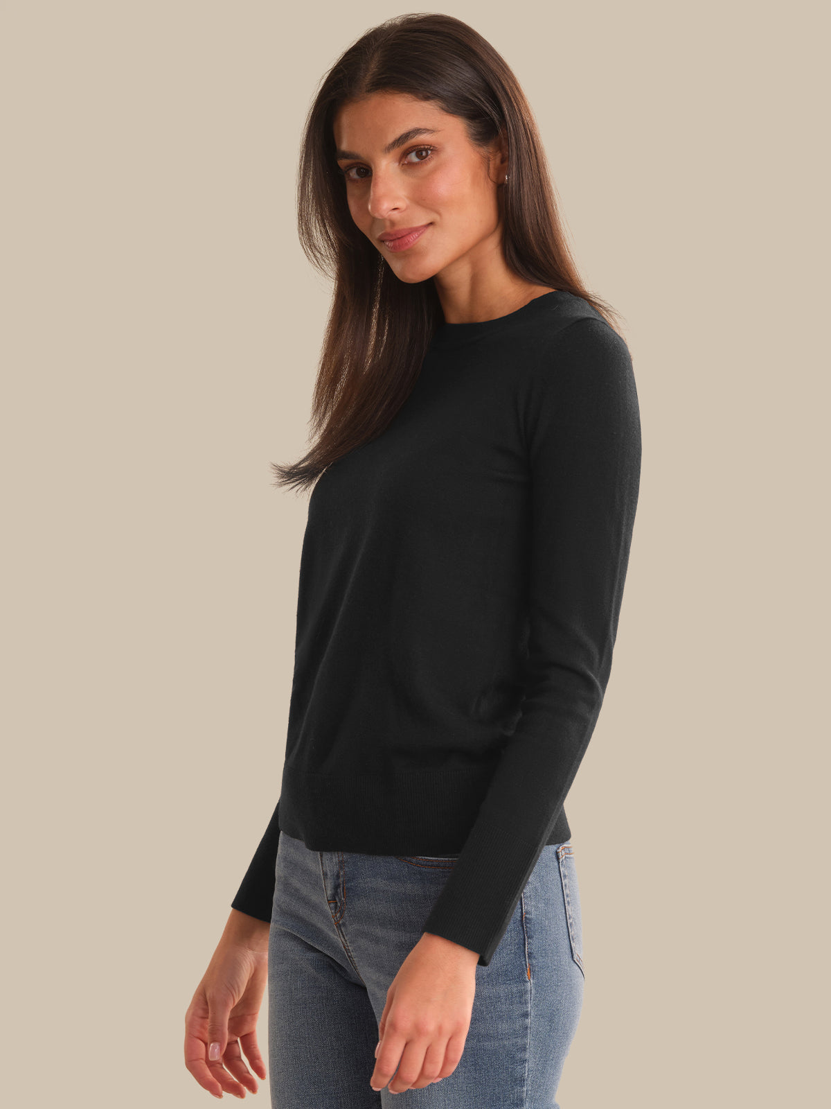A woman modeling a black long-sleeve crewneck knit top paired with blue jeans, set against a solid beige studio background.