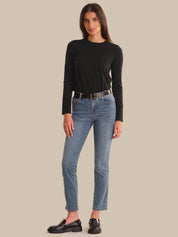 A woman modeling a black long-sleeve crewneck top, blue cropped jeans with a black belt, and black leather loafers, standing against a solid beige background.