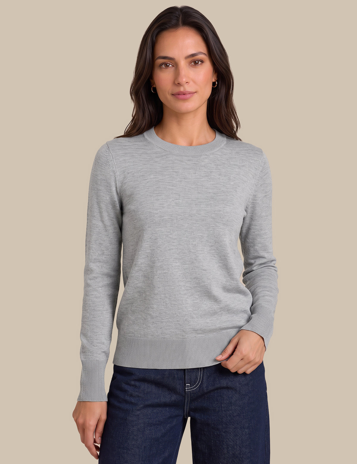 A woman modeling a light gray long-sleeve crewneck sweater with dark blue jeans, set against a solid beige background.