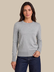 A woman modeling a light gray long-sleeve crewneck sweater with dark blue jeans, set against a solid beige background.