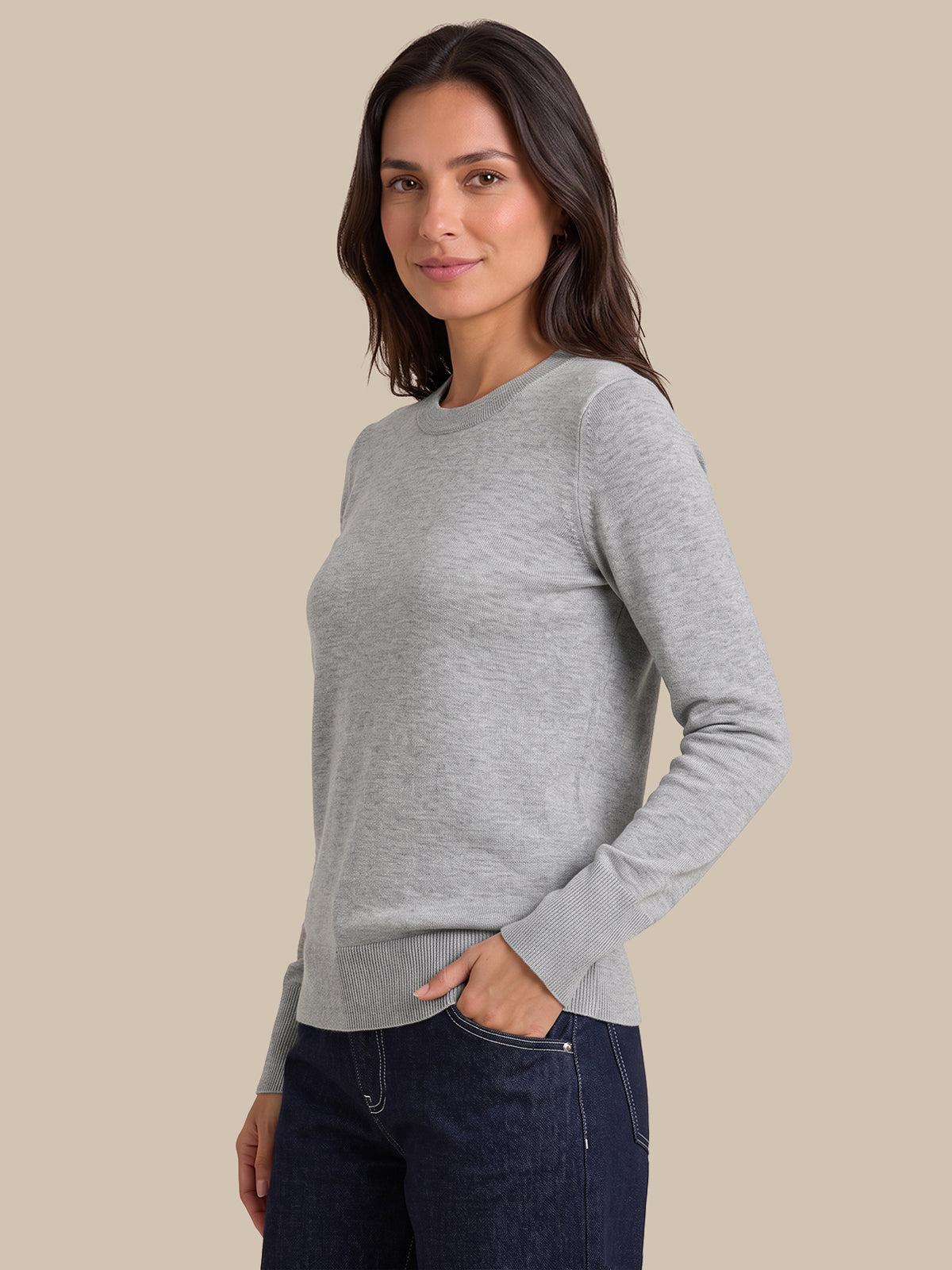 A woman modeling a light gray long-sleeve crewneck sweater, paired with dark blue jeans, against a solid beige background.