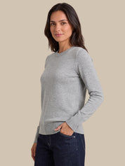 A woman modeling a light gray long-sleeve crewneck sweater, paired with dark blue jeans, against a solid beige background.