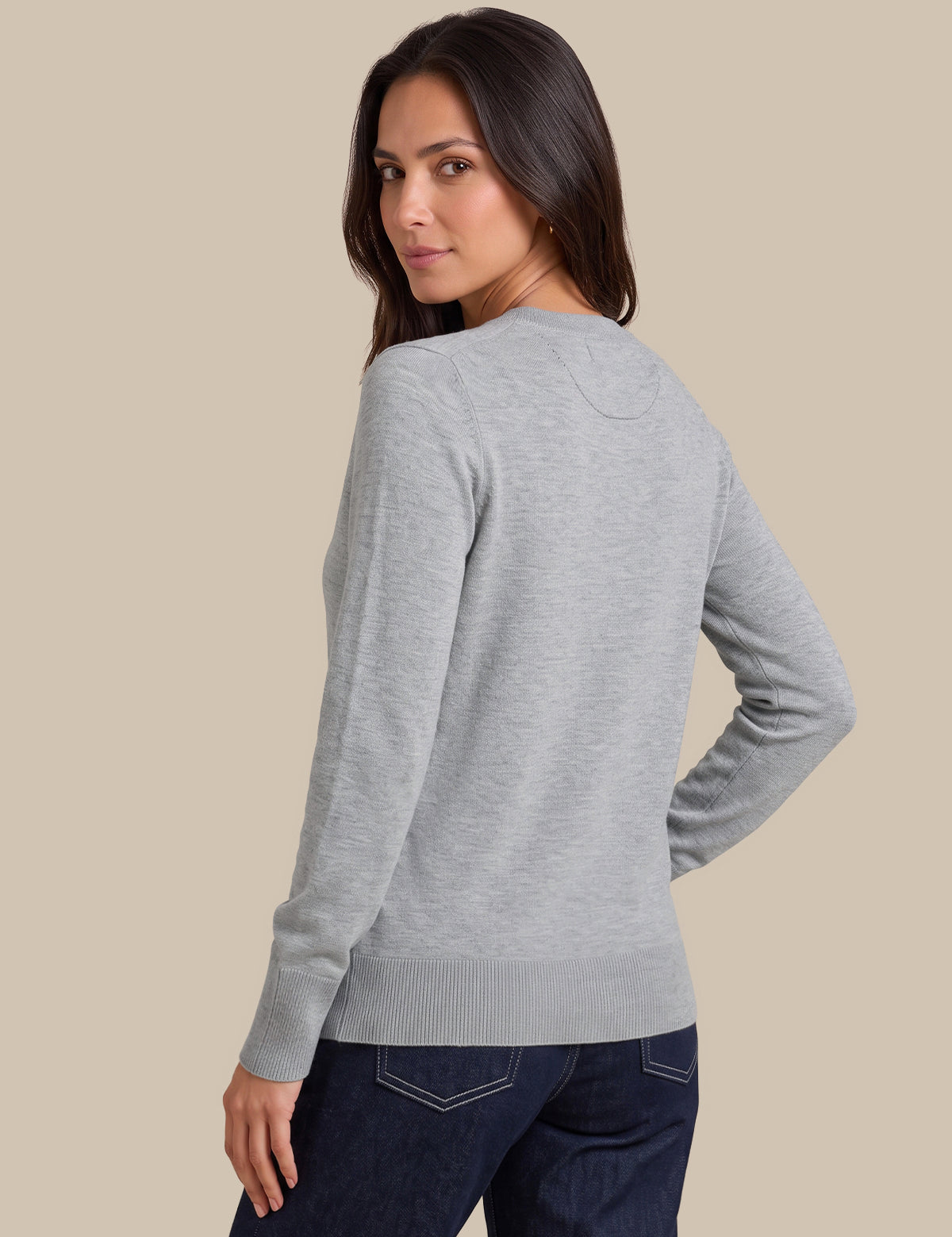 A woman wearing a light gray long-sleeve knit sweater, shown from a back-three-quarter angle against a solid beige studio background.