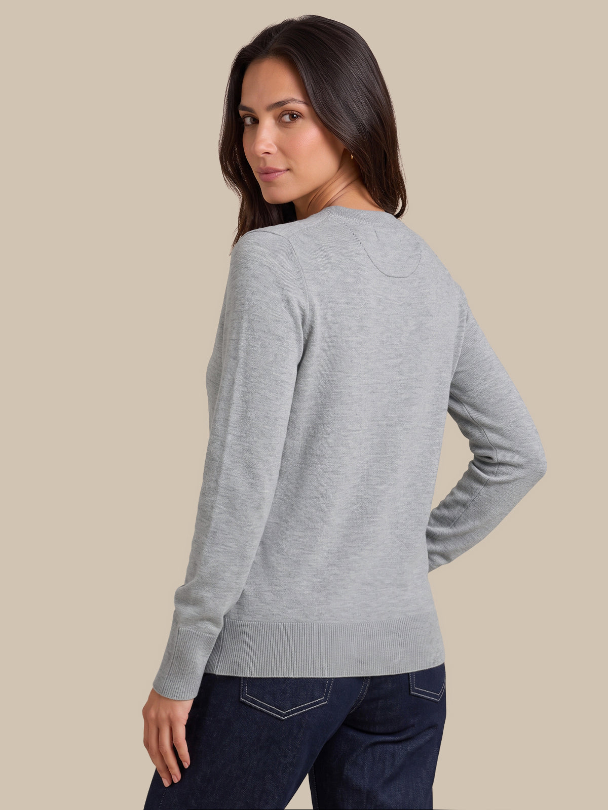 A woman wearing a light gray long-sleeve knit sweater, shown from a back-three-quarter angle against a solid beige studio background.