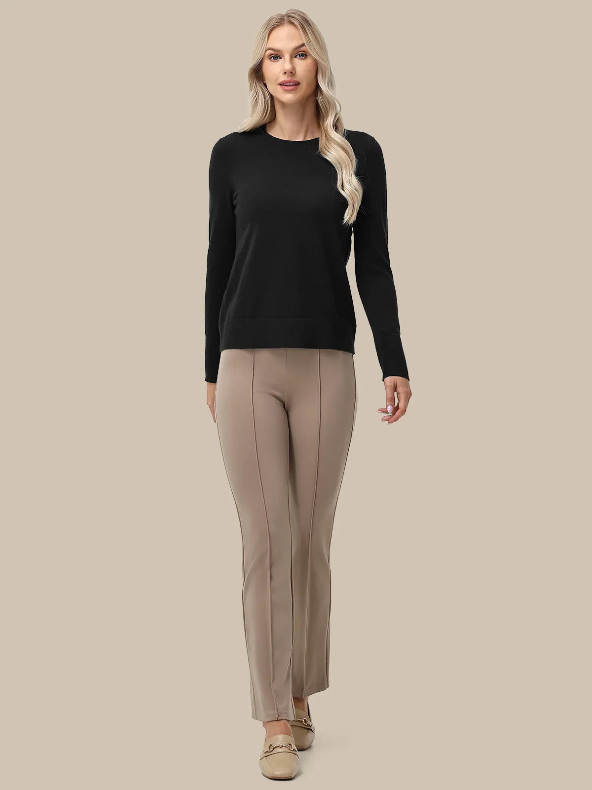 A woman modeling a black long-sleeve crewneck sweater and beige straight-leg trousers against a solid beige background.