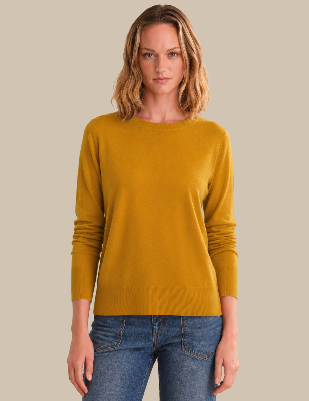 Woman wearing a mustard yellow sweater and blue jeans against a beige background