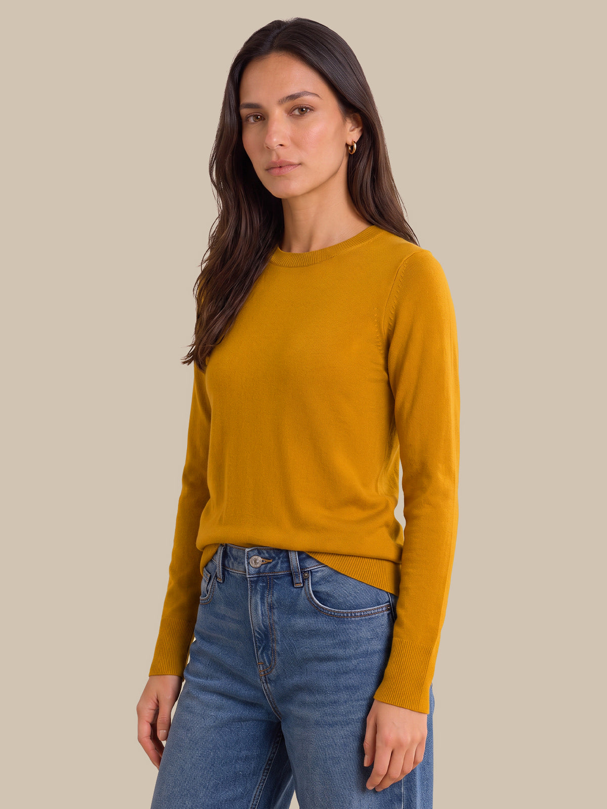 A woman modeling a mustard yellow long-sleeve crewneck sweater paired with blue jeans, set against a solid beige background.