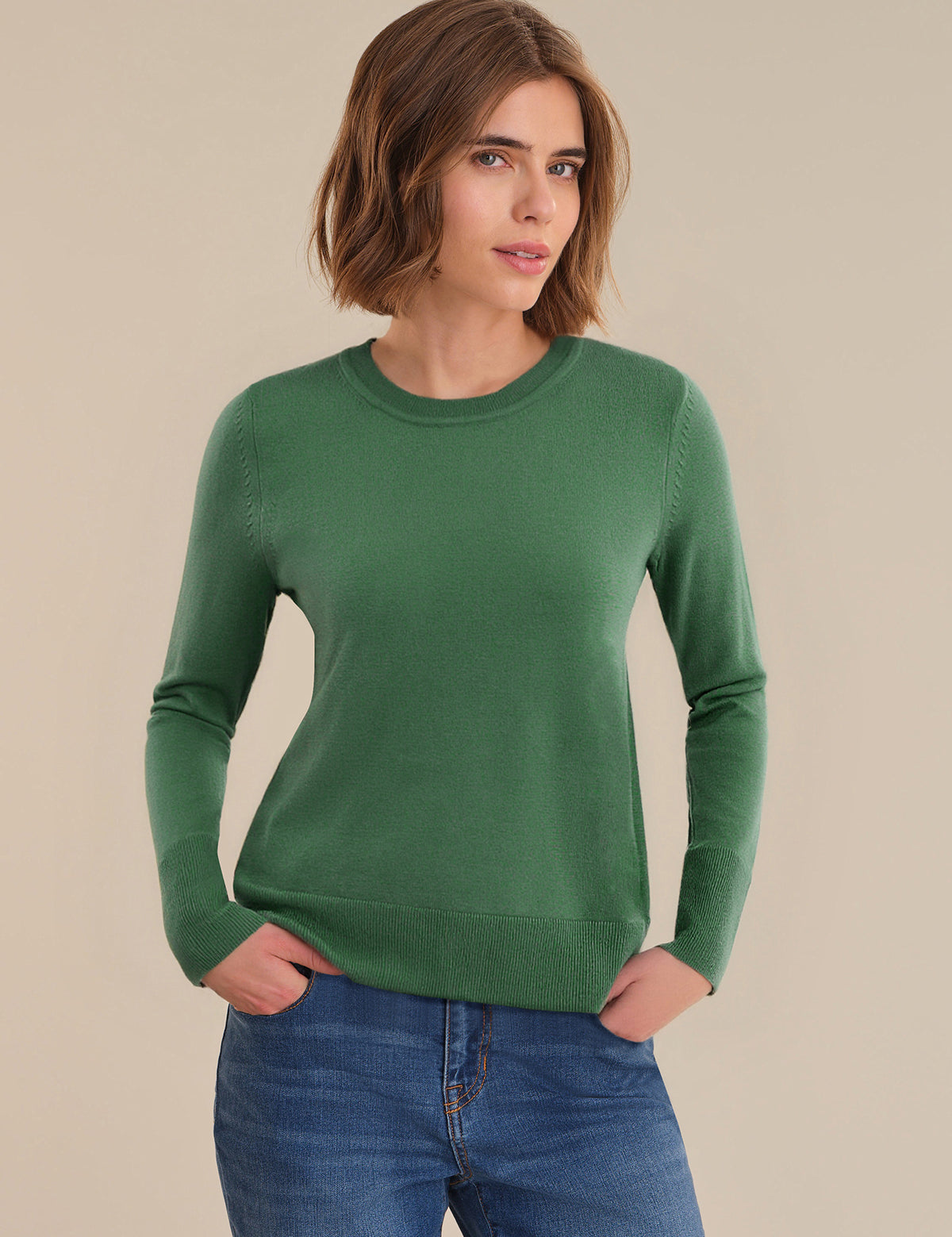 Woman wearing a green sweater and blue jeans against a beige background
