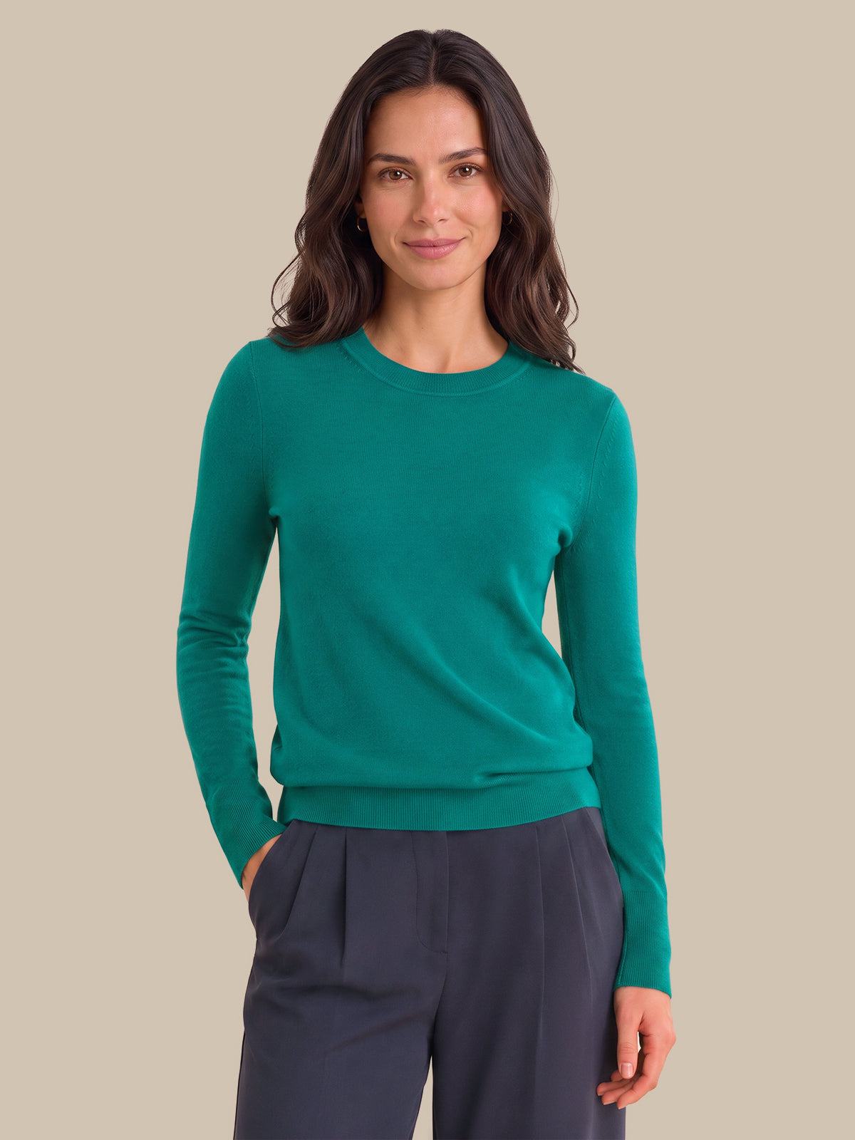 A woman modeling a long-sleeve crewneck sweater in emerald green, paired with dark gray trousers, against a solid beige background.