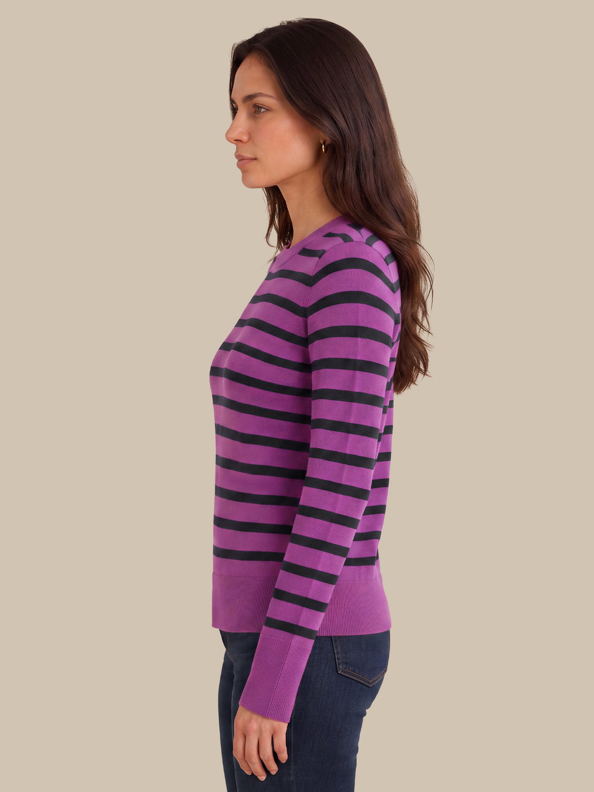 A woman shown in side profile wearing a purple and black horizontally striped long-sleeve knit sweater, paired with dark blue jeans, against a solid beige background.