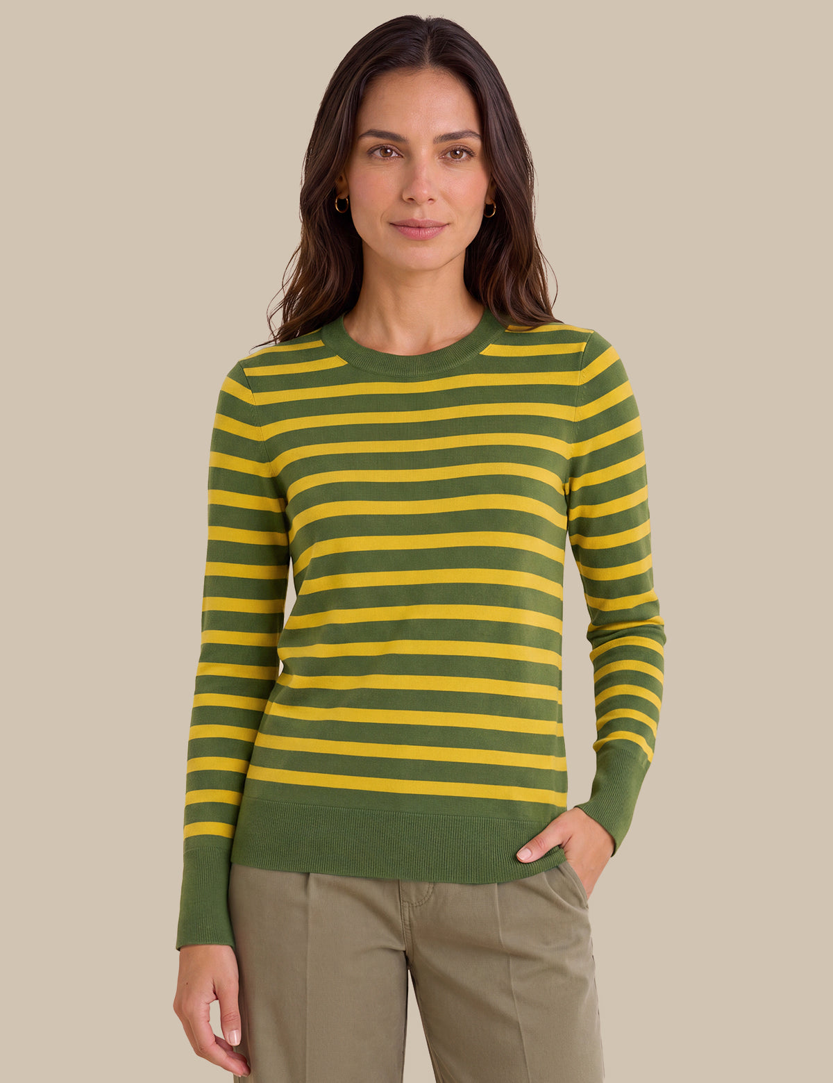 A woman modeling a green and yellow striped long-sleeve knit sweater with olive green ribbed cuffs, collar, and hem, paired with khaki pants, against a solid beige background.
