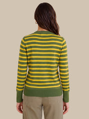 A woman viewed from the back wearing a green and yellow horizontally striped long-sleeve knit top with olive green ribbed cuffs, collar, and hem, paired with khaki pants, against a solid beige background.