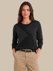 A woman modeling a black long-sleeve knit sweater with textured geometric panel details on the chest, paired with beige cargo pants, against a solid beige background.