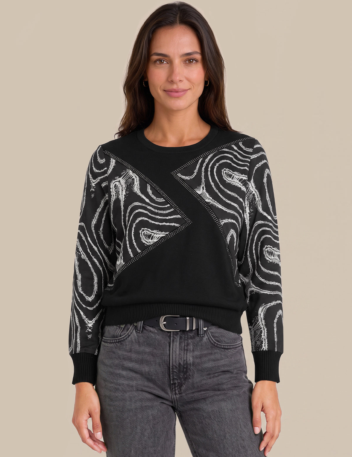 A woman modeling a black crewneck sweater with white abstract wavy patterns on the sleeves and across the chest, paired with dark gray jeans against a solid beige background.