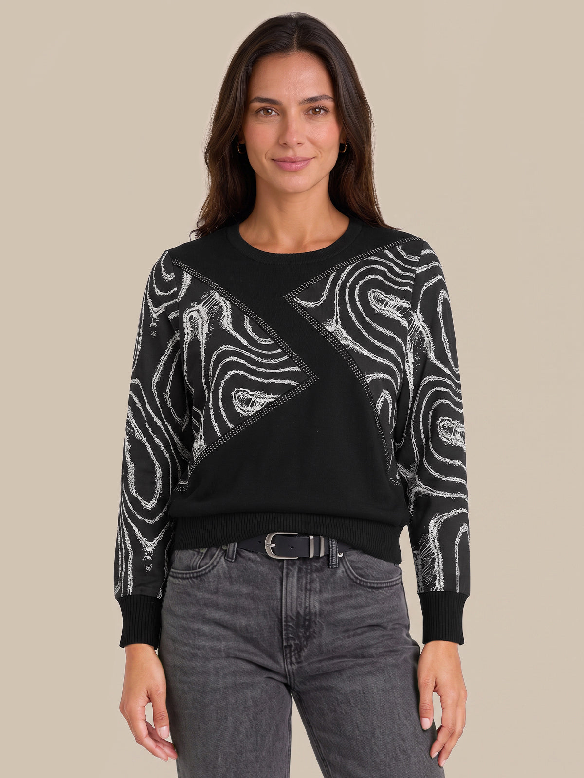 A woman modeling a black crewneck sweater with white abstract wavy patterns on the sleeves and across the chest, paired with dark gray jeans against a solid beige background.