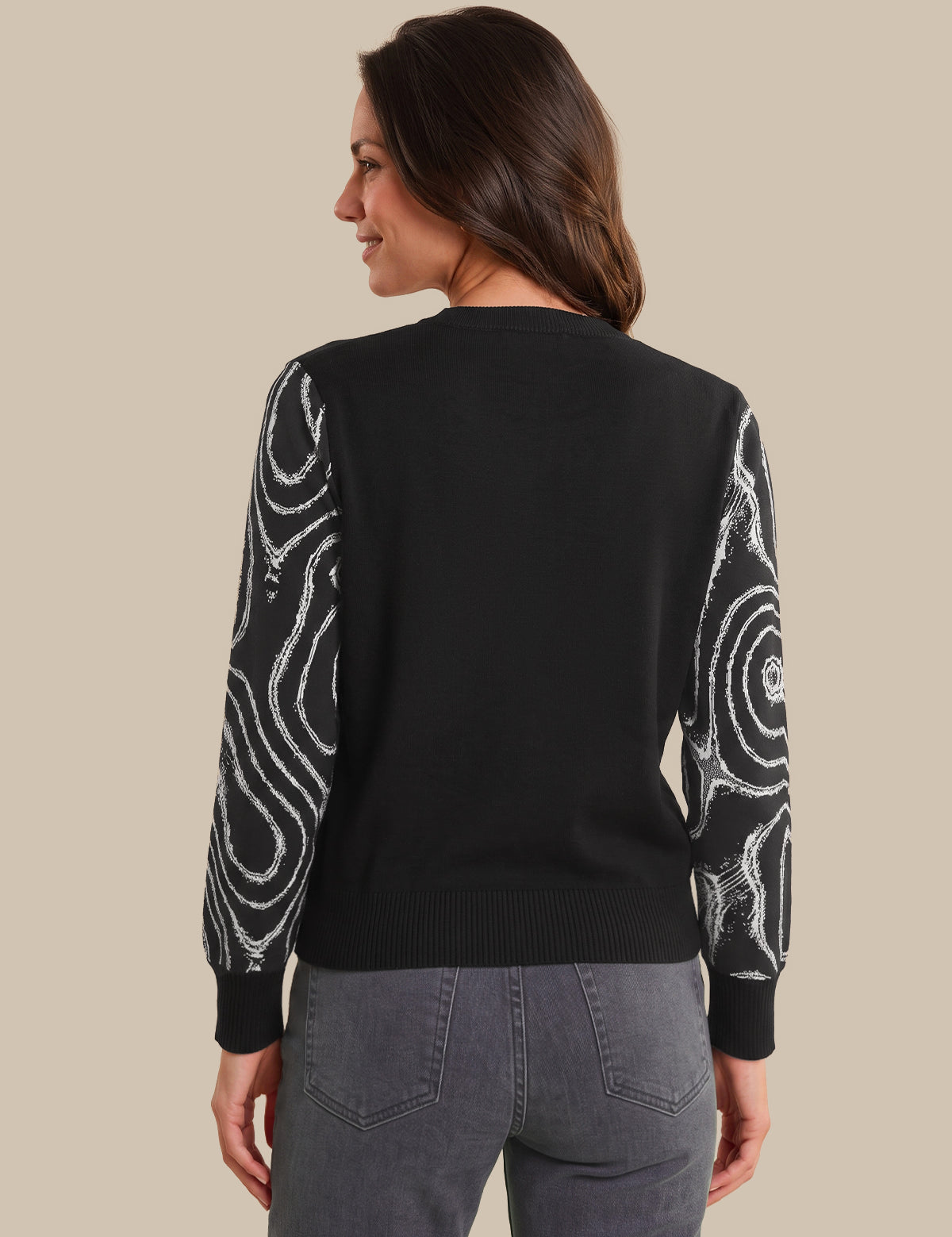 A woman shown from the back wearing a black long-sleeve sweater with white swirling abstract patterns on the sleeves, paired with gray jeans, against a solid beige background.