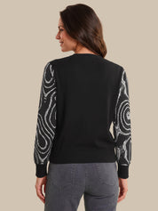 A woman shown from the back wearing a black long-sleeve sweater with white swirling abstract patterns on the sleeves, paired with gray jeans, against a solid beige background.