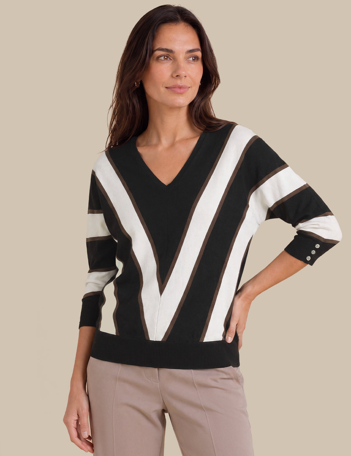 A woman wearing a black and white striped V-neck sweater with three-quarter sleeves and beige trousers, standing against a solid beige background.