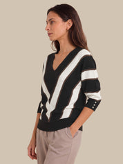 A woman wearing a black and white striped V-neck sweater with three-button cuffs, paired with light gray trousers, shown in profile against a solid beige background.