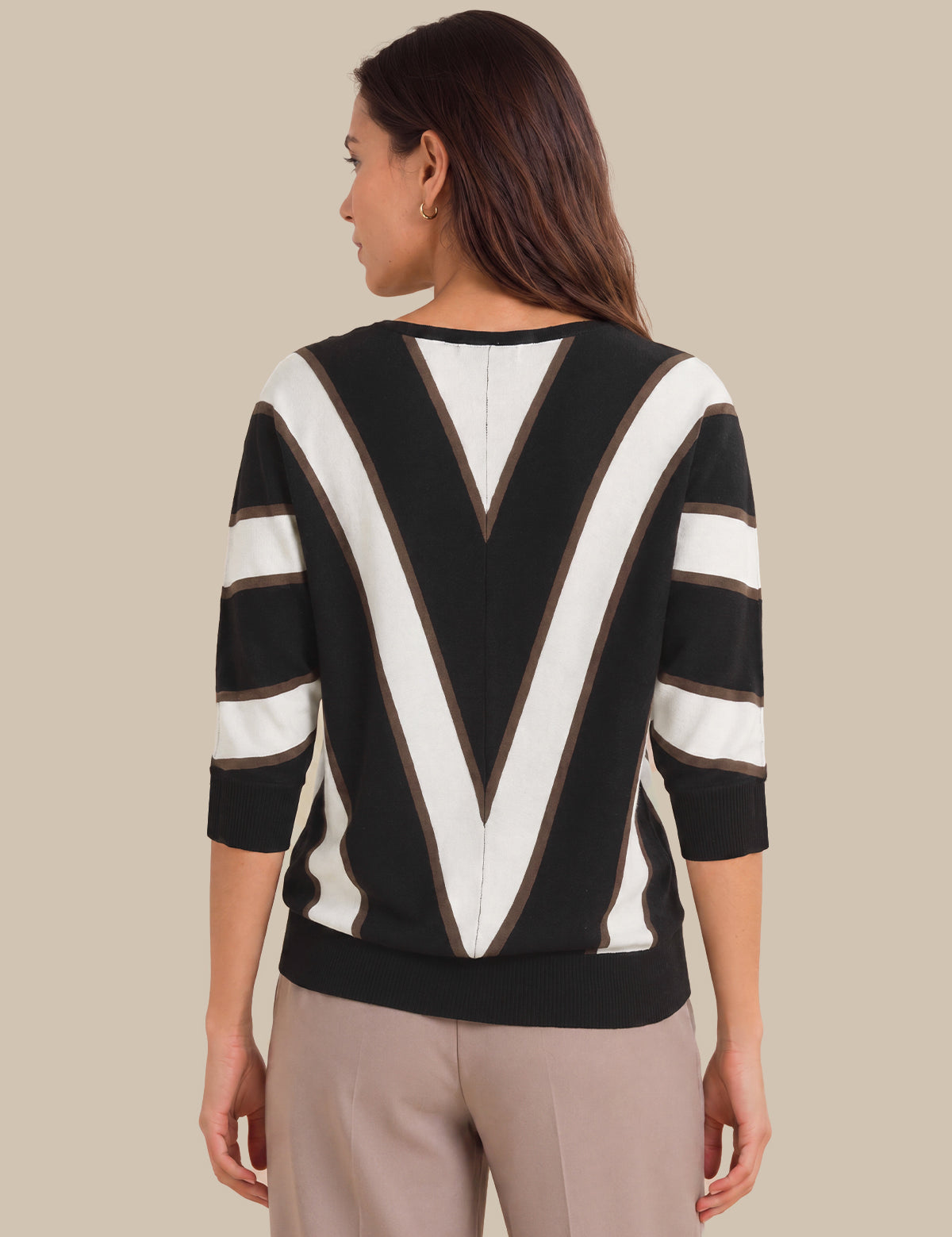 A woman shown from the back wearing a black and white striped sweater with a large V-shaped stripe pattern on the back and three-quarter sleeves, paired with beige trousers, against a solid beige background.