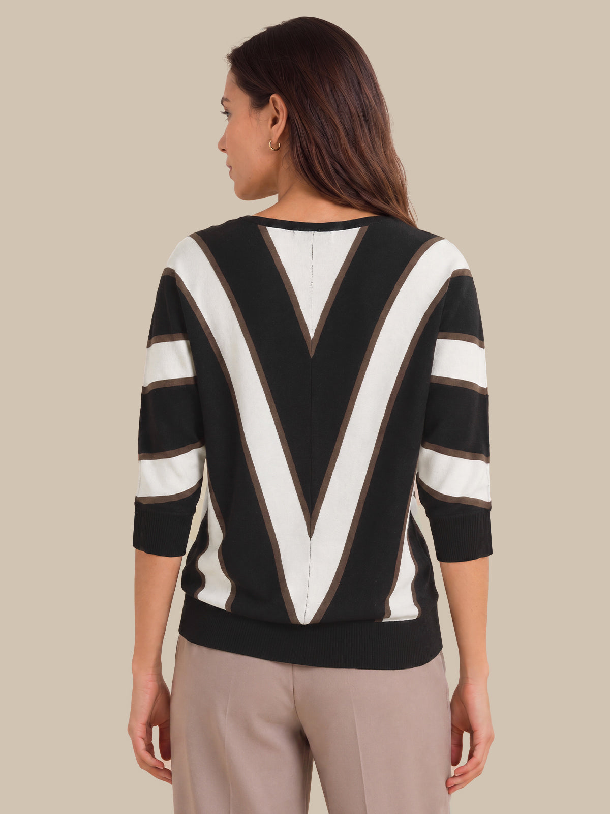 A woman shown from the back wearing a black and white striped sweater with a large V-shaped stripe pattern on the back and three-quarter sleeves, paired with beige trousers, against a solid beige background.