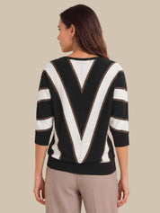 A woman shown from the back wearing a black and white striped sweater with a large V-shaped stripe pattern on the back and three-quarter sleeves, paired with beige trousers, against a solid beige background.