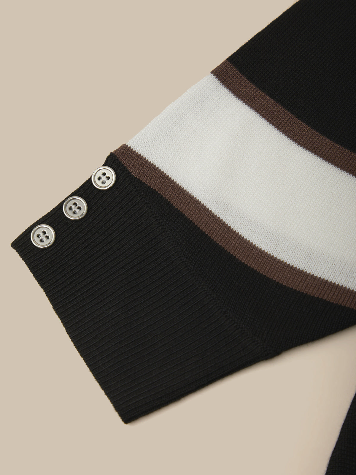 Close-up of a ribbed knit sweater sleeve in black, white, and brown stripes, featuring three decorative buttons on the cuff, set against a solid beige background.
