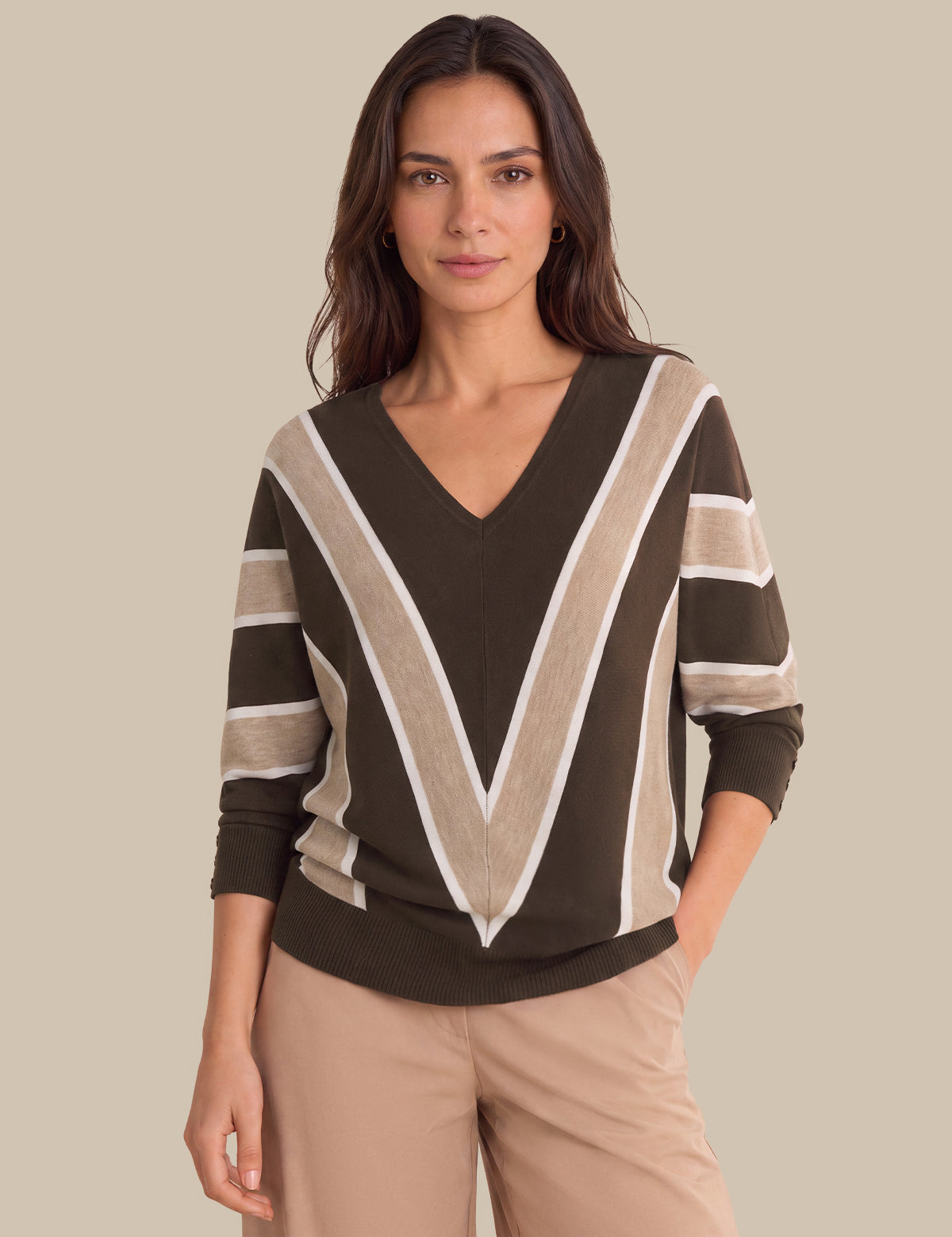 A woman modeling a brown V-neck sweater with beige and white striped accents on the shoulders and sleeves, paired with beige pants, against a solid beige background.
