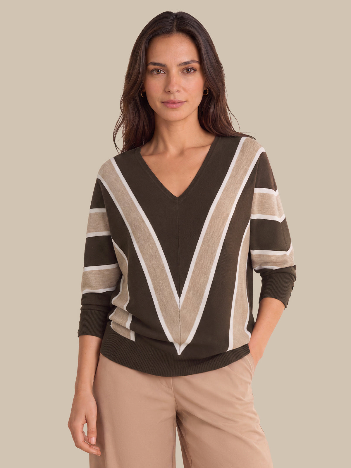 A woman modeling a brown V-neck sweater with beige and white striped accents on the shoulders and sleeves, paired with beige pants, against a solid beige background.