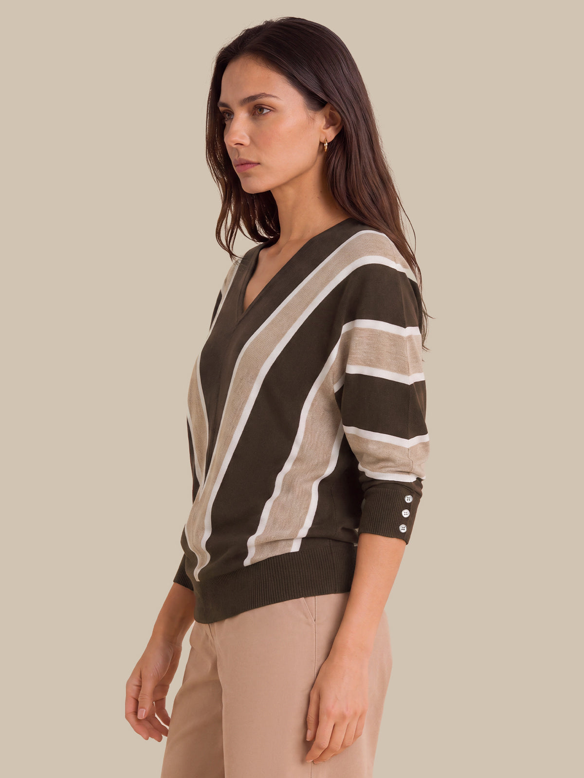 A woman wearing a V-neck knit sweater with diagonal stripes in olive green, beige, and white, paired with beige trousers, set against a solid beige background.