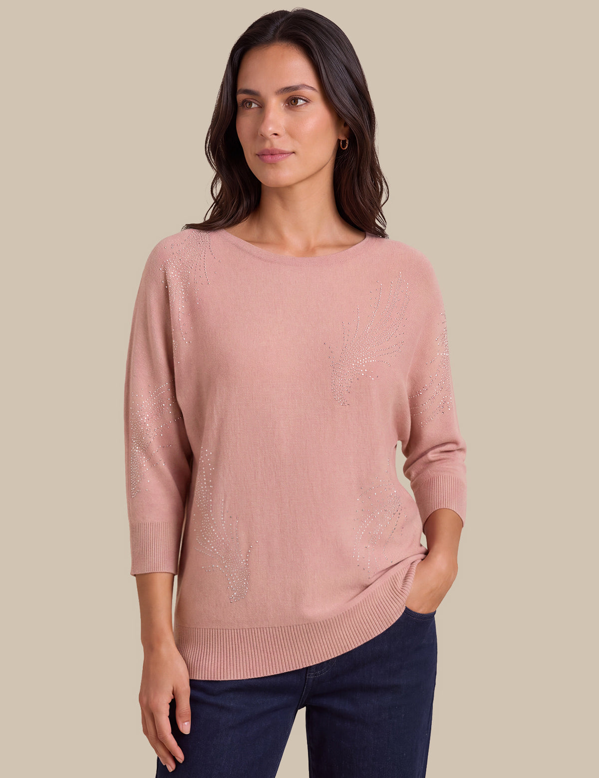 A woman modeling a dusty pink knit sweater with subtle tonal floral embroidery, paired with dark blue jeans, against a solid beige background.