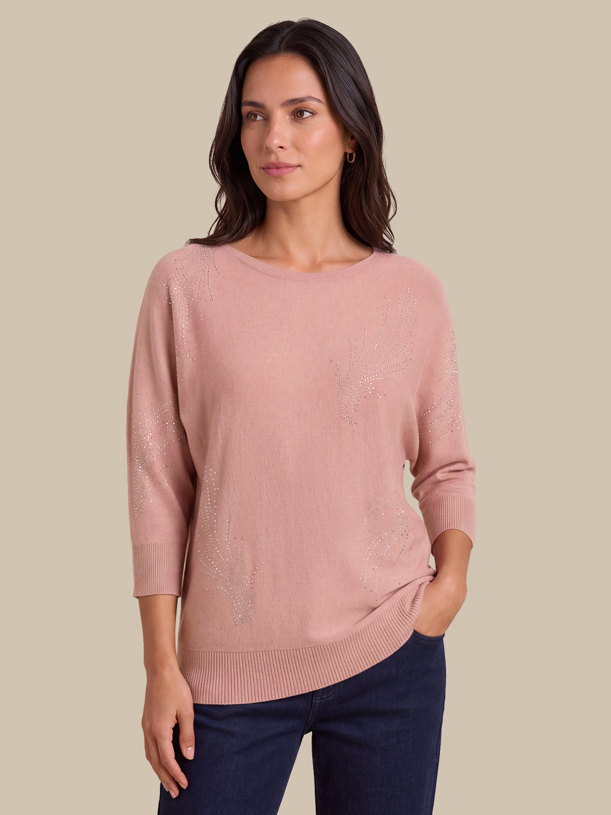A woman modeling a dusty pink knit sweater with subtle tonal floral embroidery, paired with dark blue jeans, against a solid beige background.