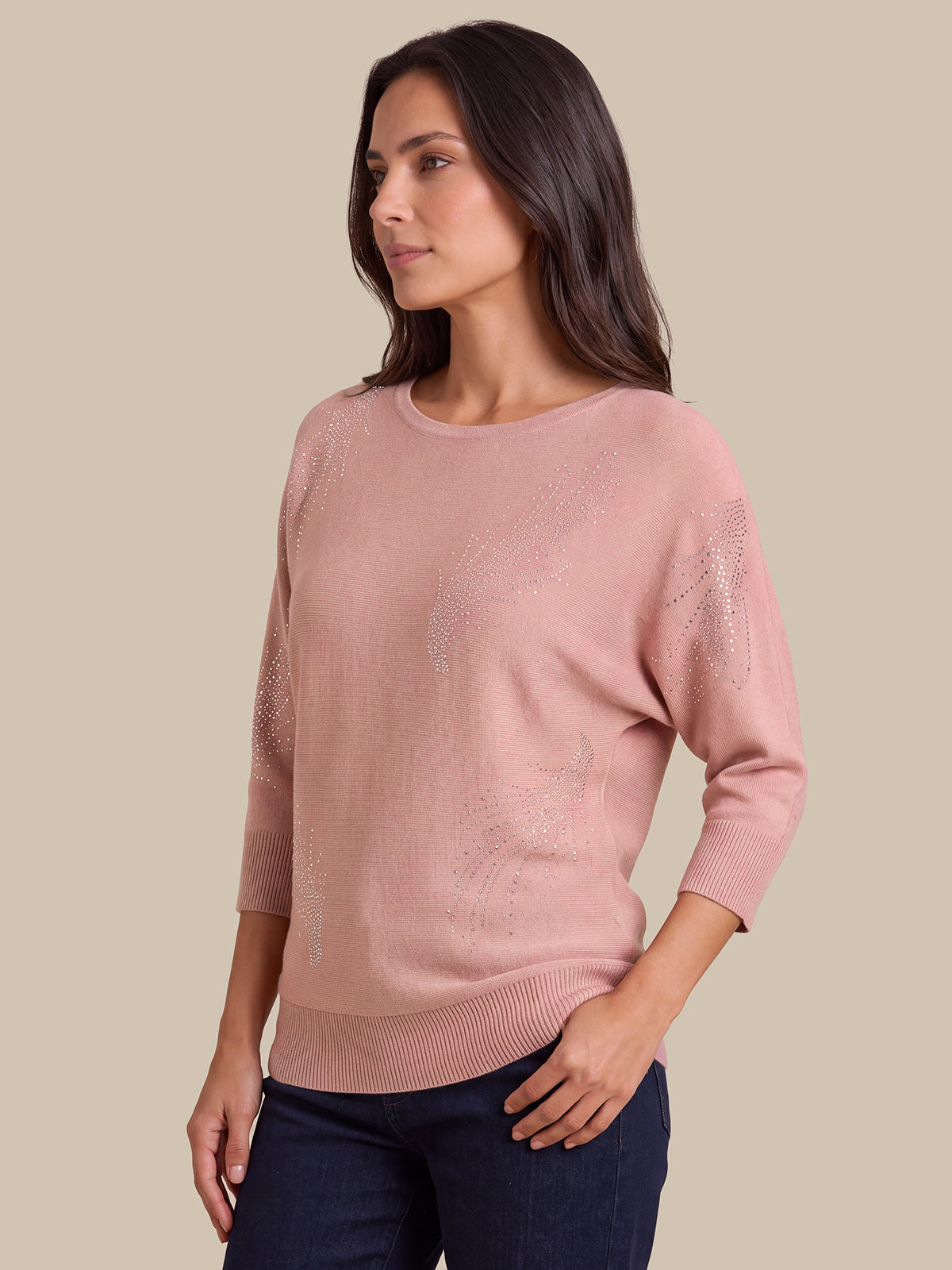 A woman wearing a dusty pink knit sweater with subtle textured patterns on the front and sleeves, paired with dark blue jeans, set against a solid beige background.