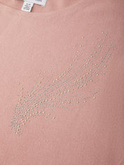 Close-up of a dusty pink knit top with a rhinestone embellishment forming a stylized feather or wing shape on the chest.
