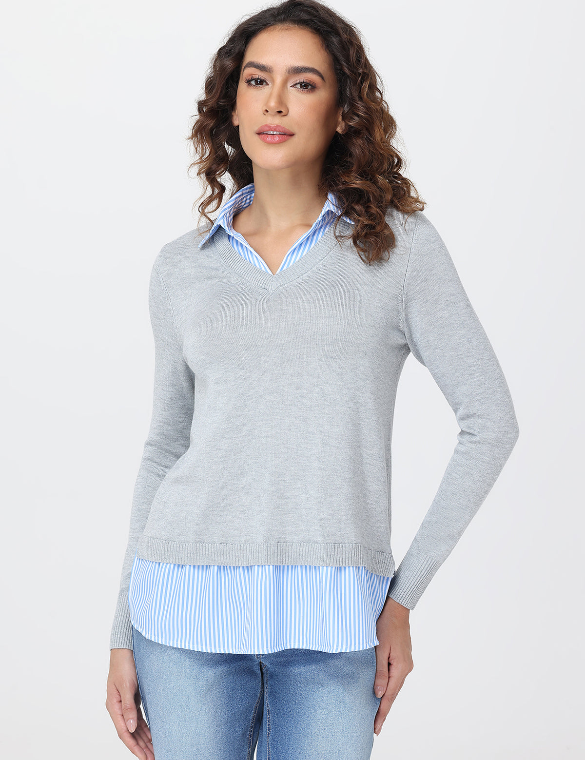 Woman wearing a gray sweater over a striped shirt with blue jeans on a white background