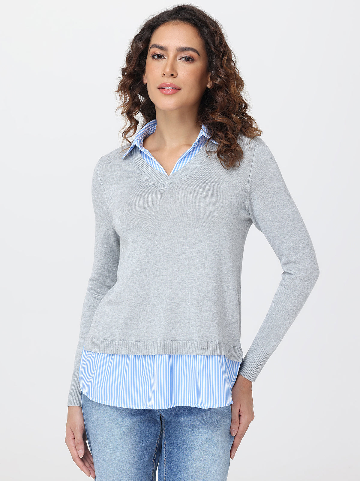Woman wearing a gray sweater over a striped shirt with blue jeans on a white background