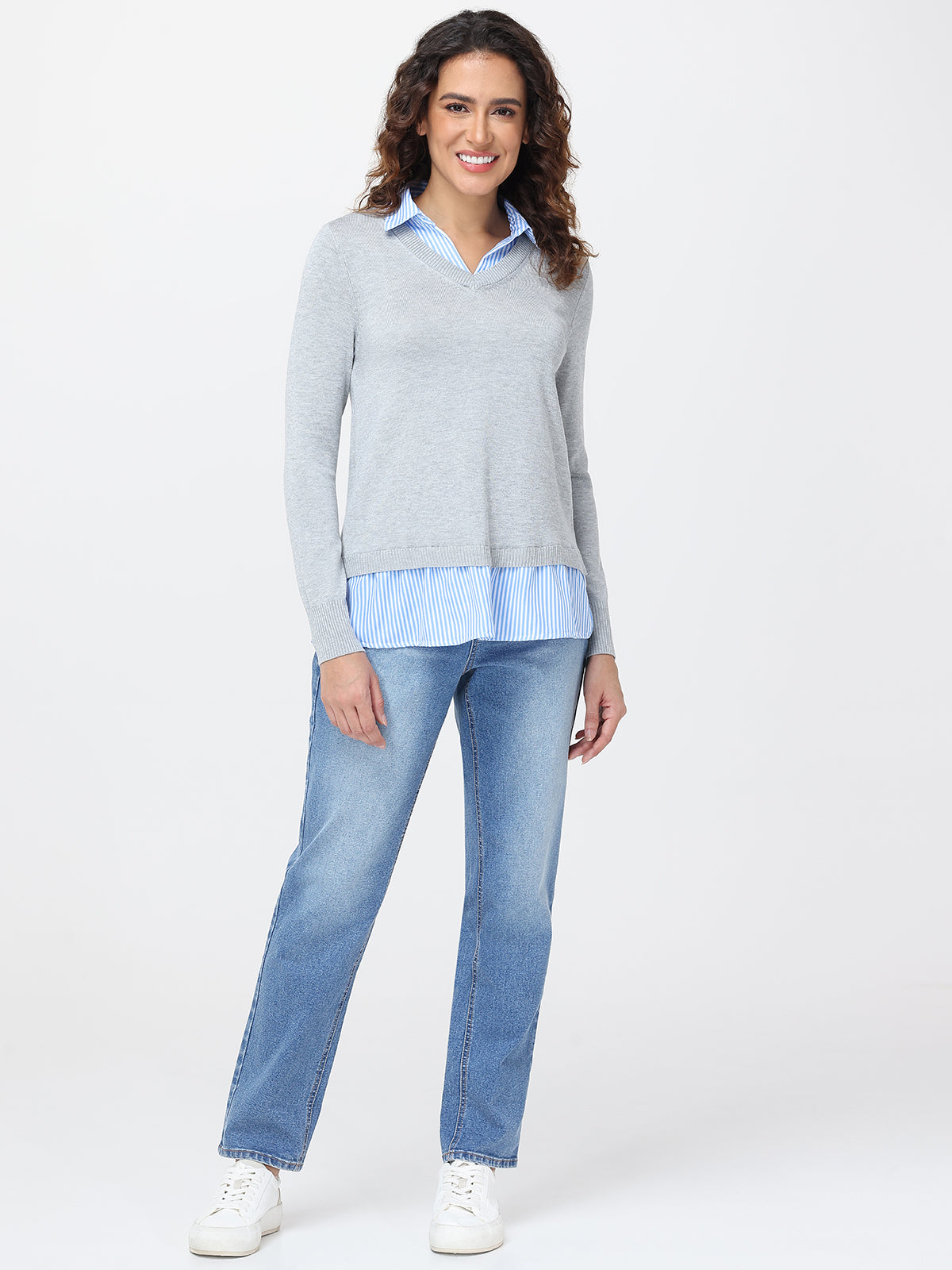 Woman wearing a light gray sweater over a blue shirt with jeans on a white background