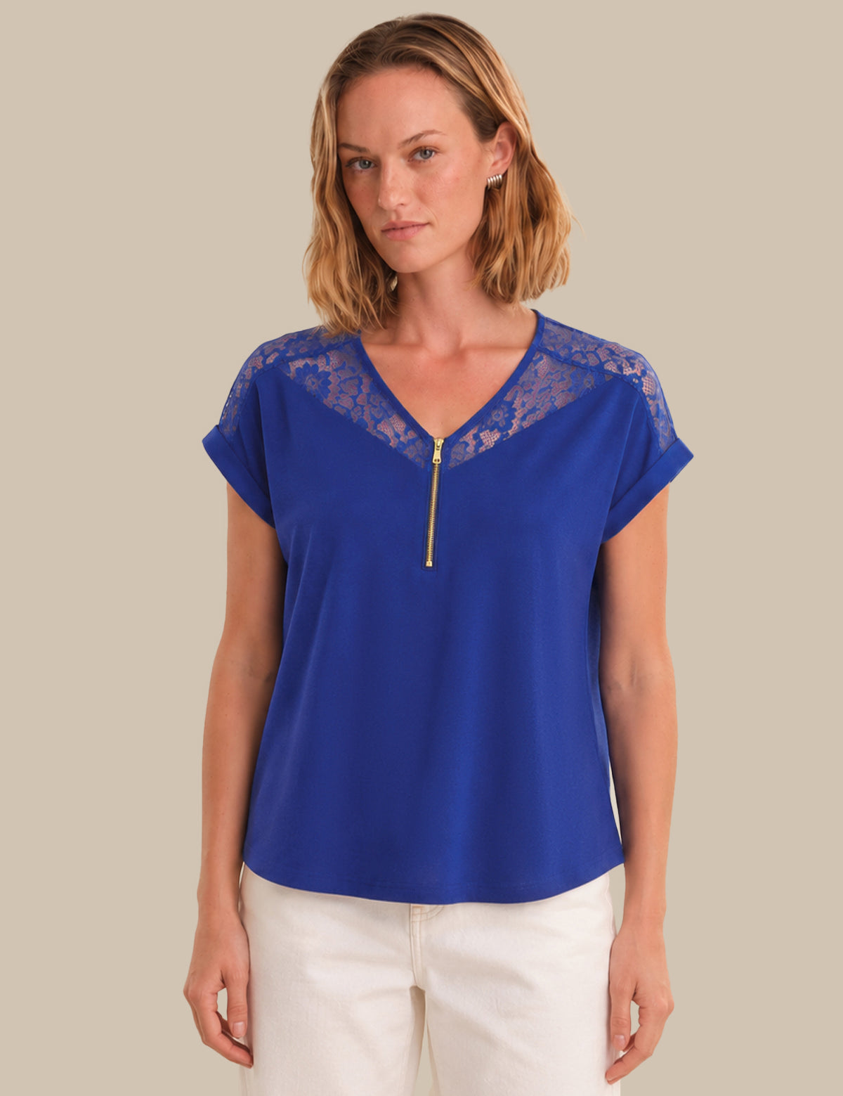 A woman modeling a royal blue short-sleeve top with a lace V-neckline and a gold zipper detail, paired with white pants, against a solid beige background.