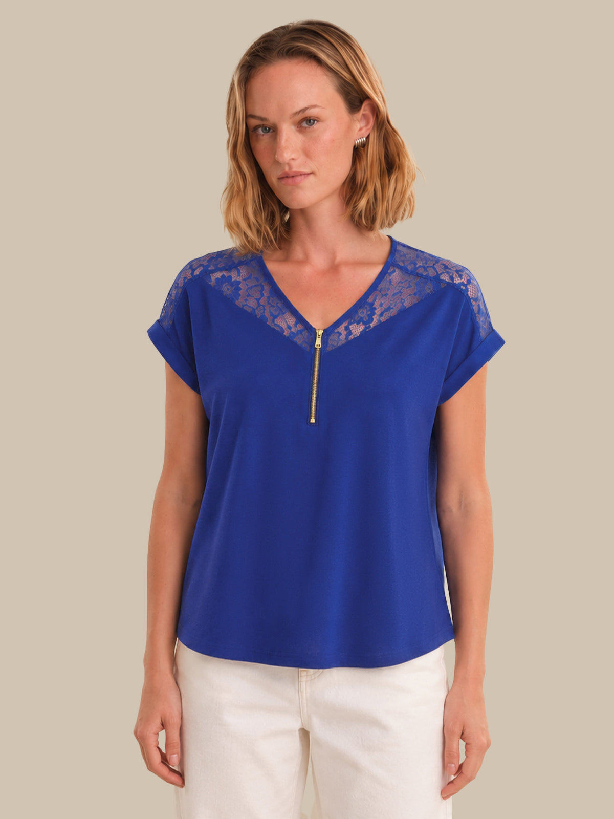 A woman modeling a royal blue short-sleeve top with a lace V-neckline and a gold zipper detail, paired with white pants, against a solid beige background.