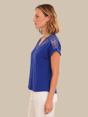 A woman shown in profile wearing a royal blue short-sleeve top with sheer lace detailing on the shoulders and a front zipper, paired with white pants, against a solid beige background.