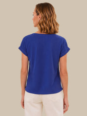 A woman shown from the back wearing a royal blue short-sleeve top and cream-colored pants, set against a solid beige studio background.